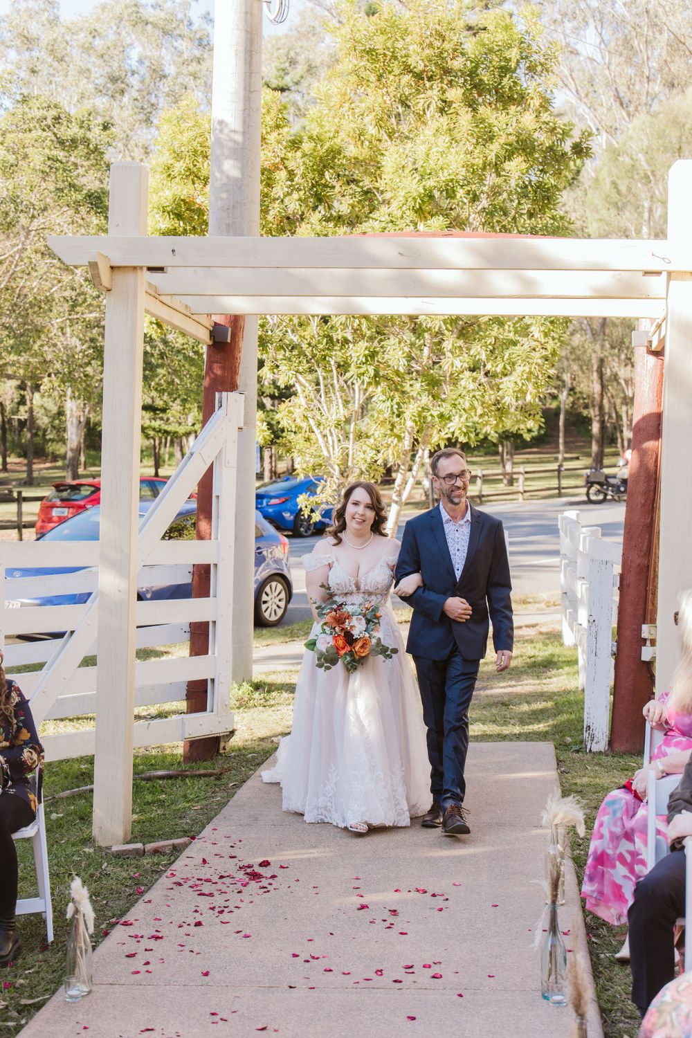 Wedding party members process down an outdoor walkway decorated with white posts and rose petals.