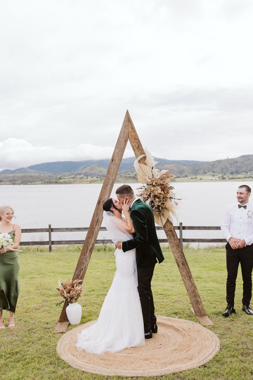 Wooden triangle wedding arch decorated with dried flowers stands against lakeside mountain backdrop.