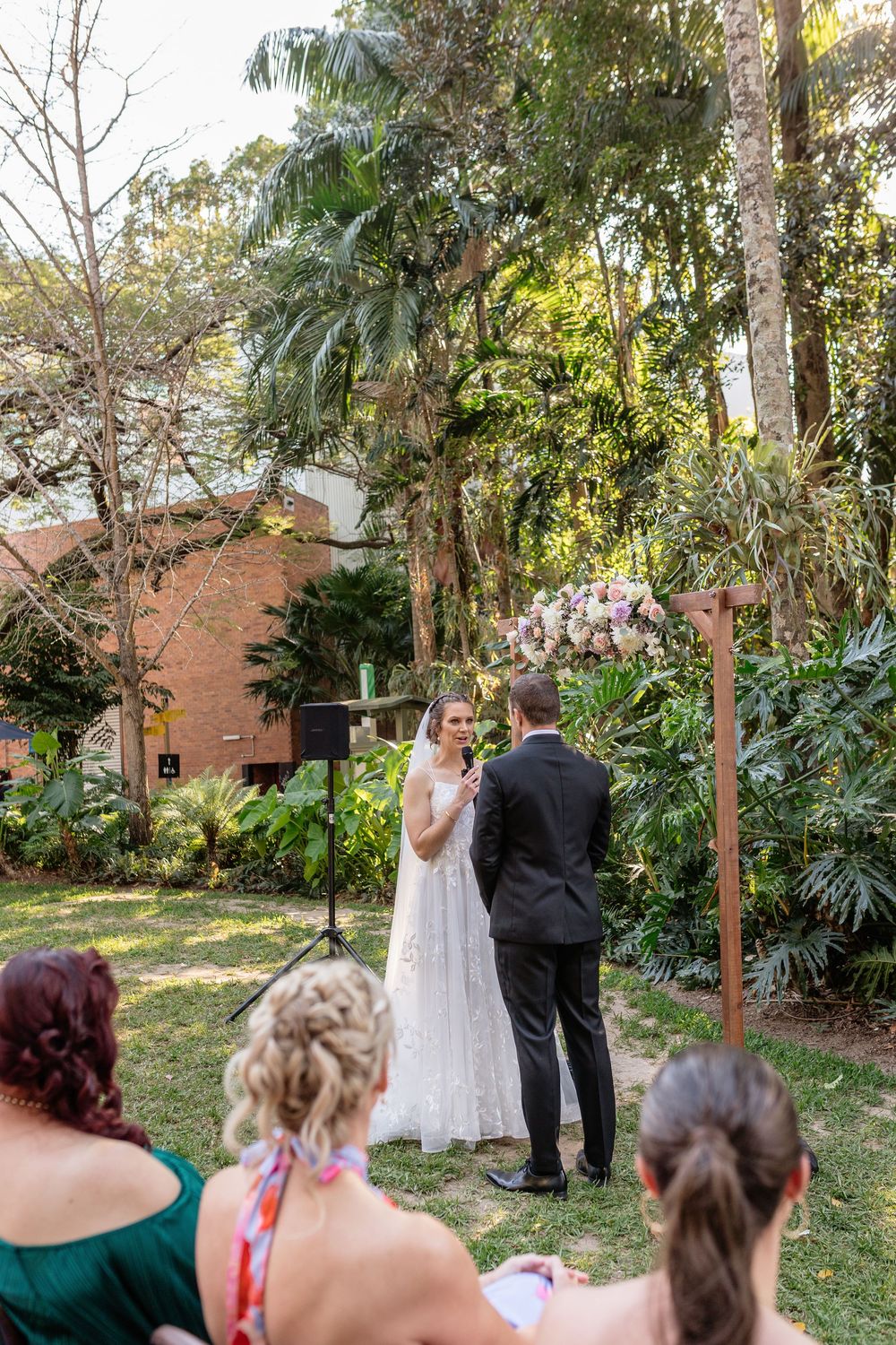 Wedding ceremony taking place in a lush garden setting with guests seated and watching the couple exchange vows.