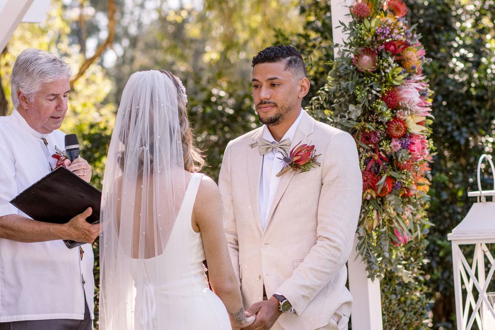 A romantic outdoor garden wedding ceremony with people gathered under floral decorations and white gazebo.