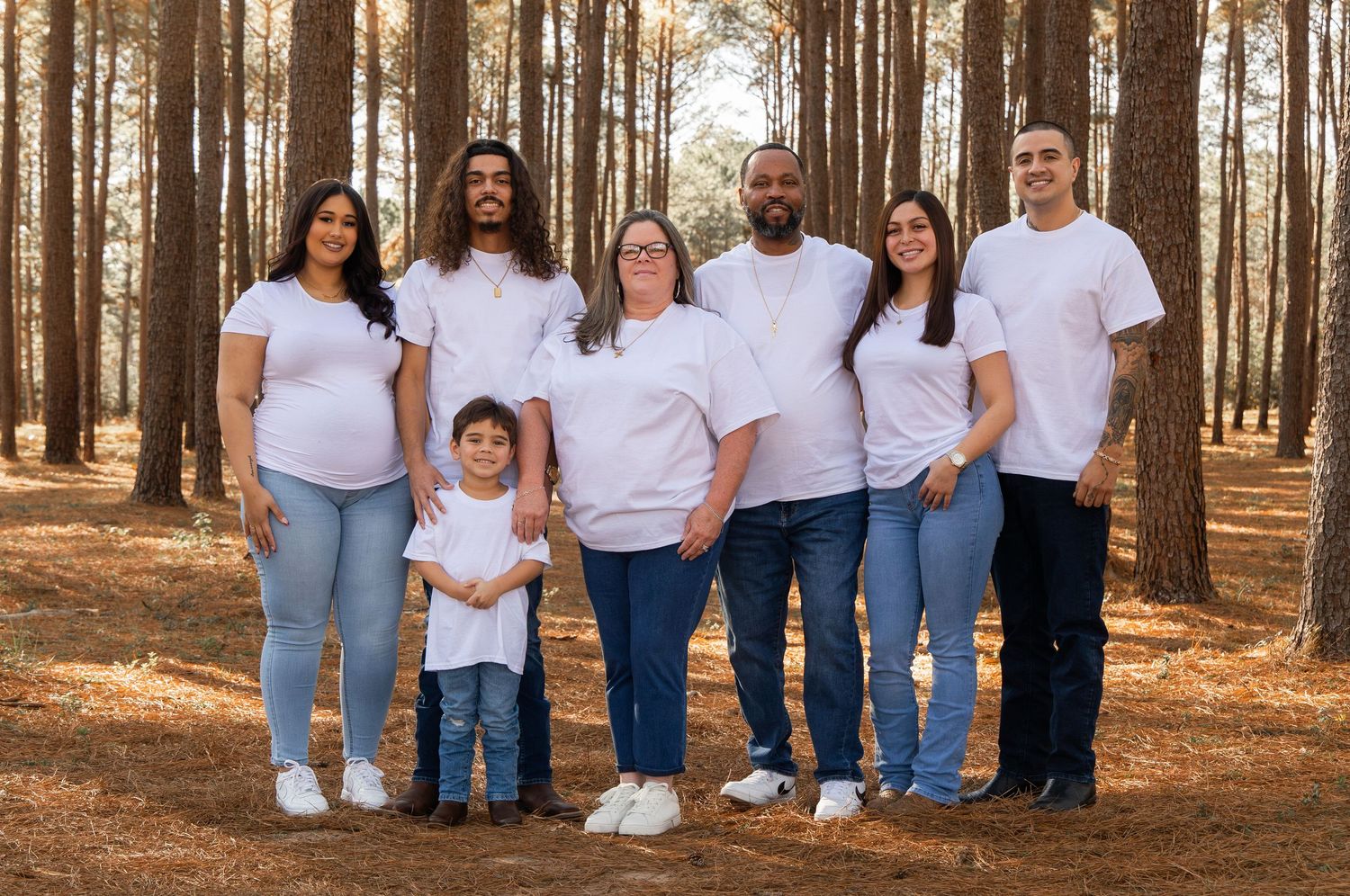 Family group wearing coordinated white shirts and blue jeans poses together in a sunlit pine forest setting.