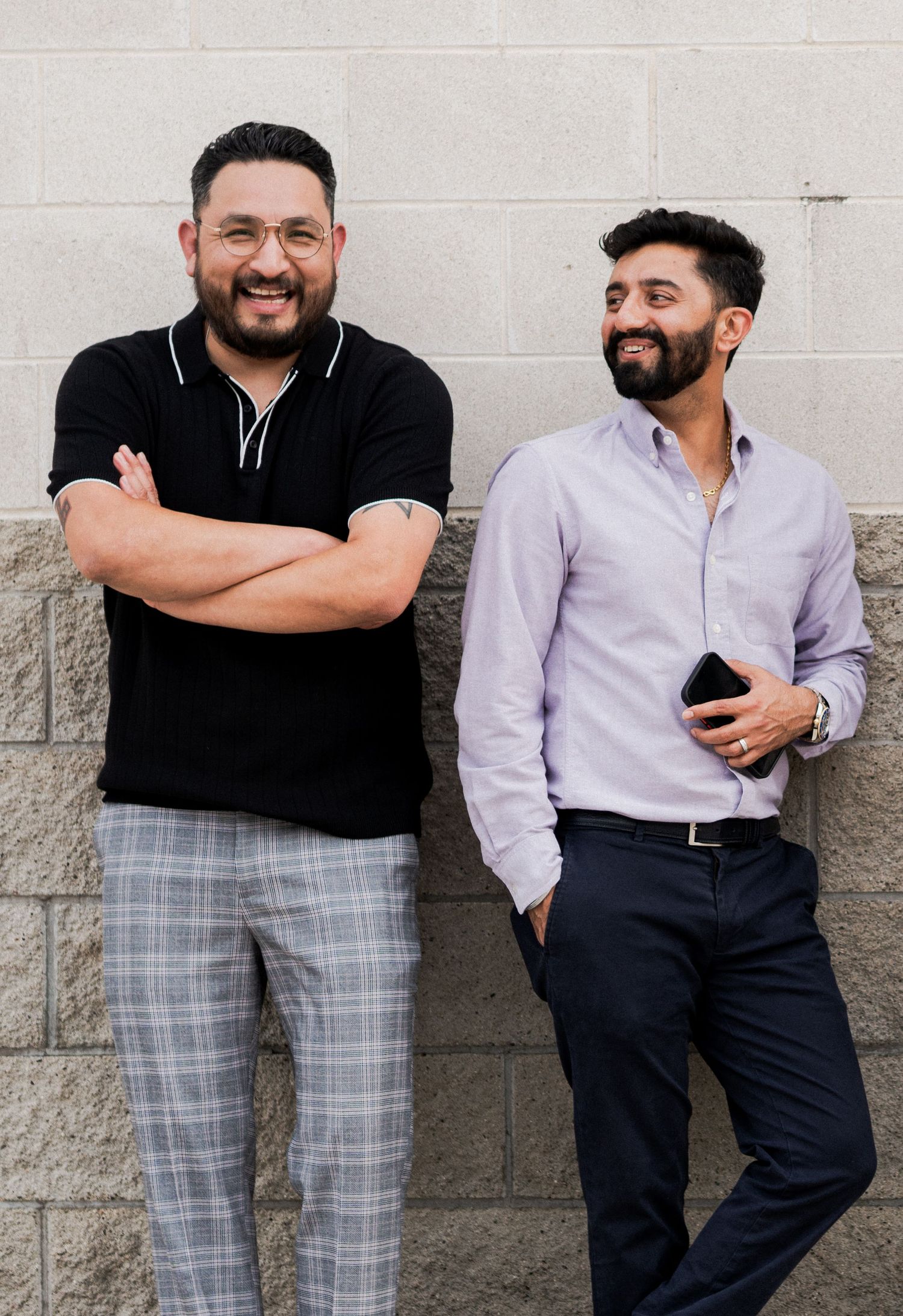 Two colleagues in business casual attire pose against a gray wall with confident smiles.