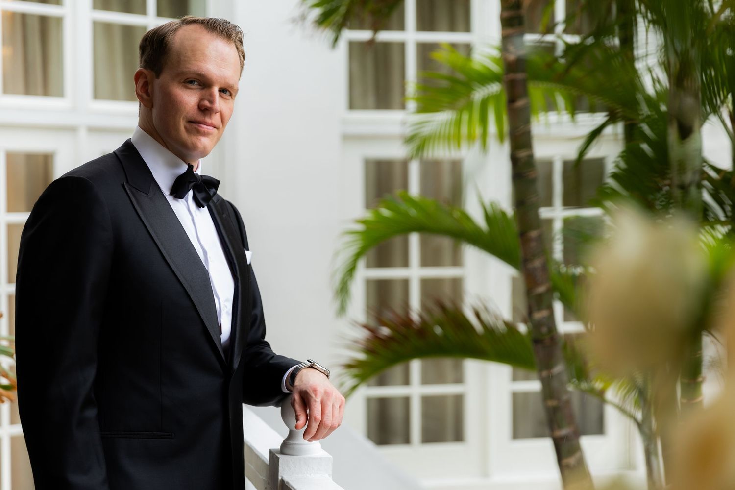 Elegant groom portrait in classic black tuxedo against white tropical resort background.