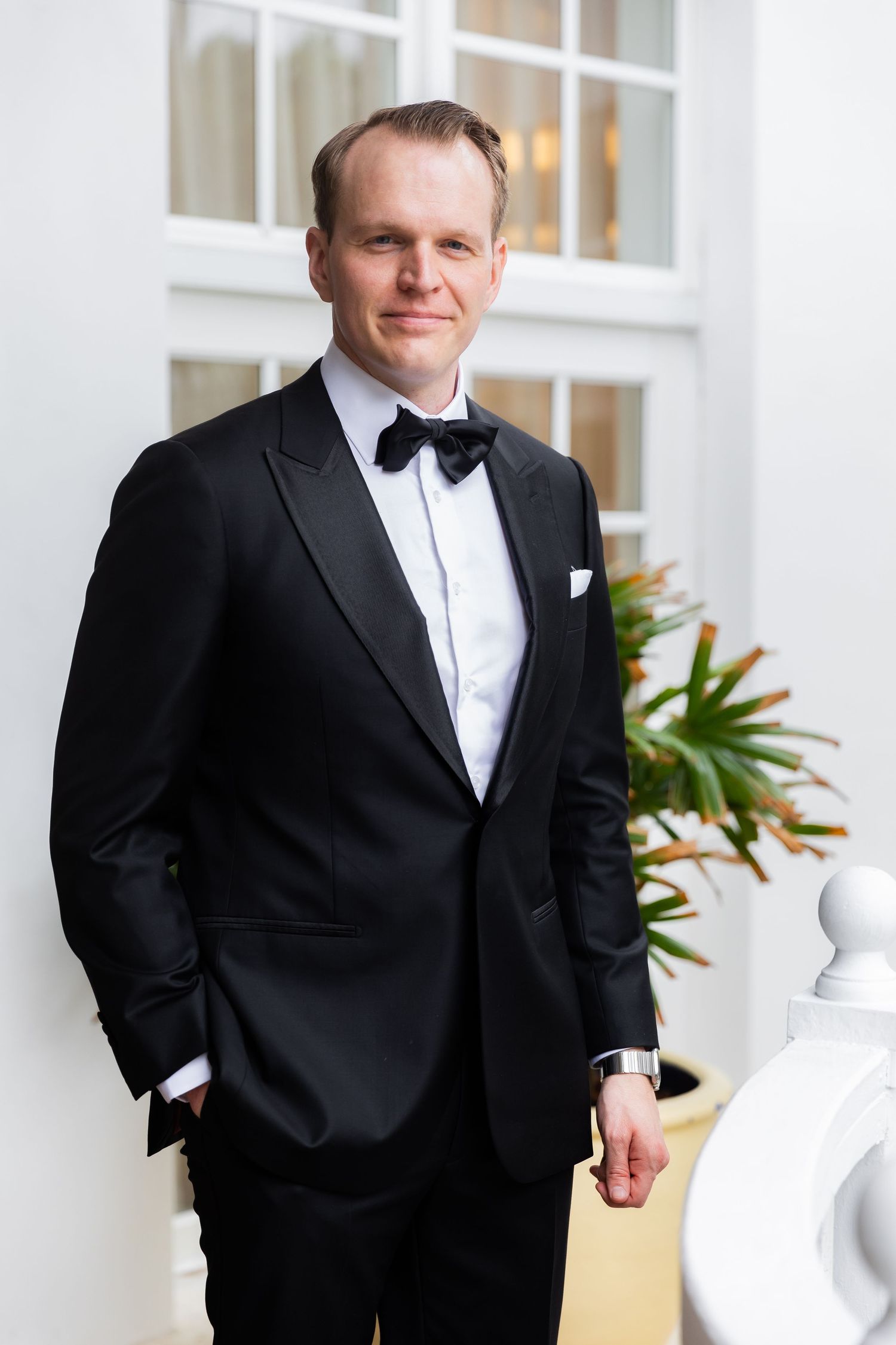 Man in classic black tuxedo with bowtie poses against white interior wall.