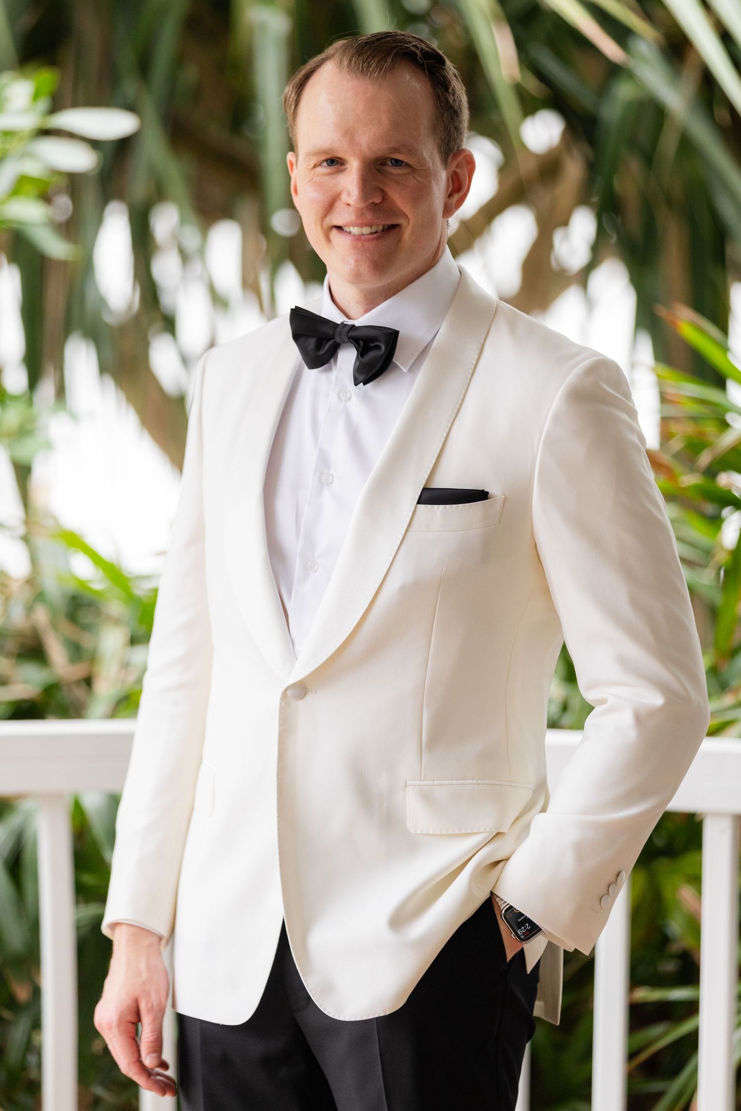 Groom in white dinner jacket and black bow tie poses on tropical veranda.