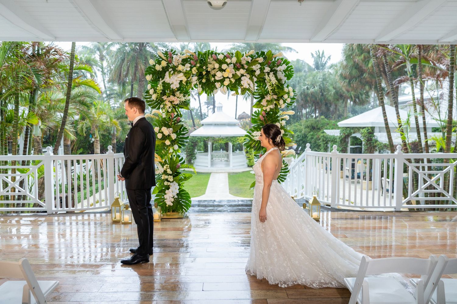 Wedding ceremony setup on covered porch with floral arch and white chairs in rainy weather.