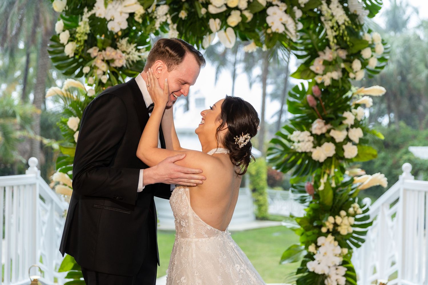Couple embraces during wedding ceremony under luxurious white floral arch installation.