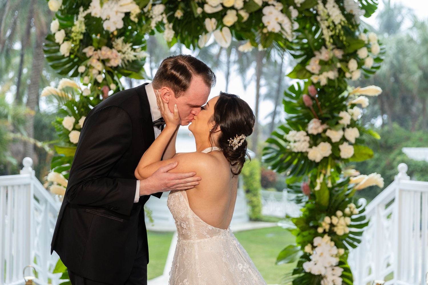 Romantic moment captured under elaborate white flower arch during outdoor wedding ceremony.