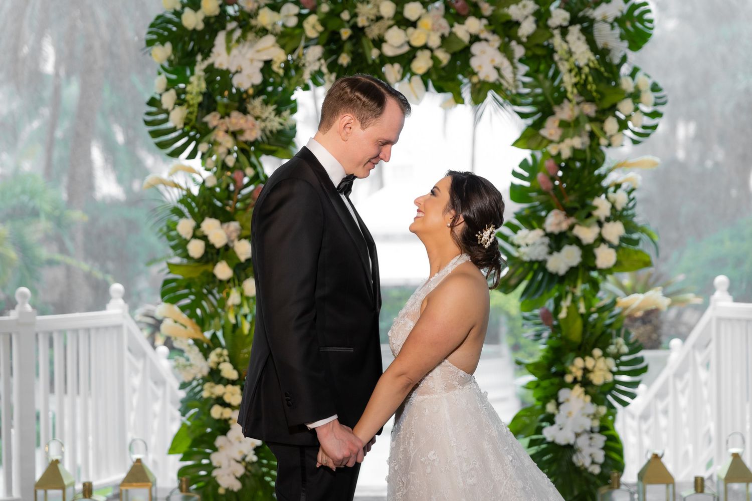Intimate wedding ceremony moment under lush white floral arch with palm trees in background.