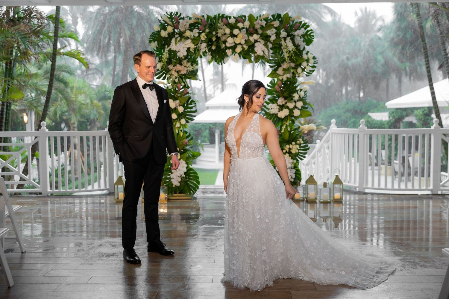 Bride in flowing white gown poses with groom under elaborate floral arch during rainy ceremony.