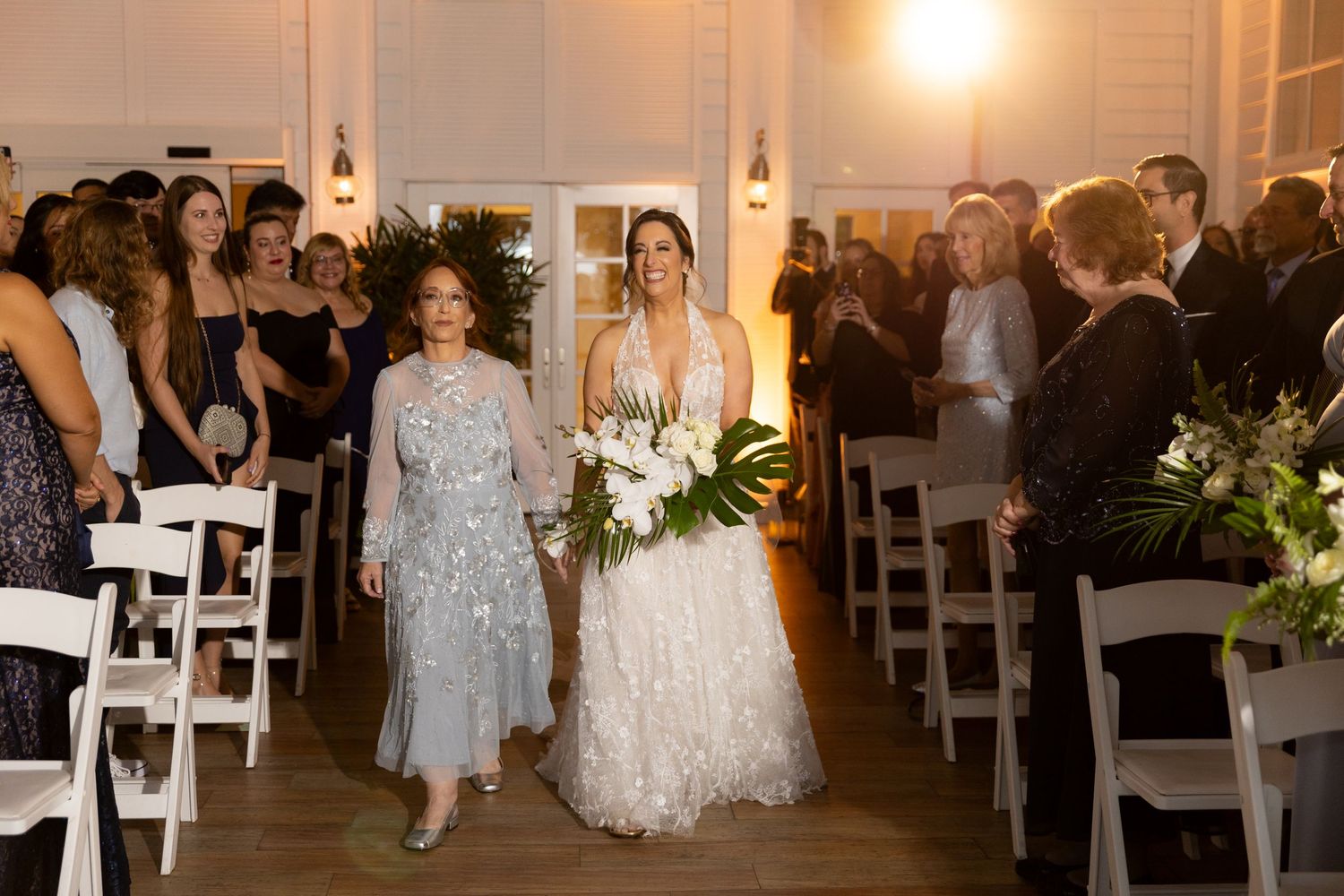Wedding ceremony in a bright indoor venue with white chairs and floral decorations.