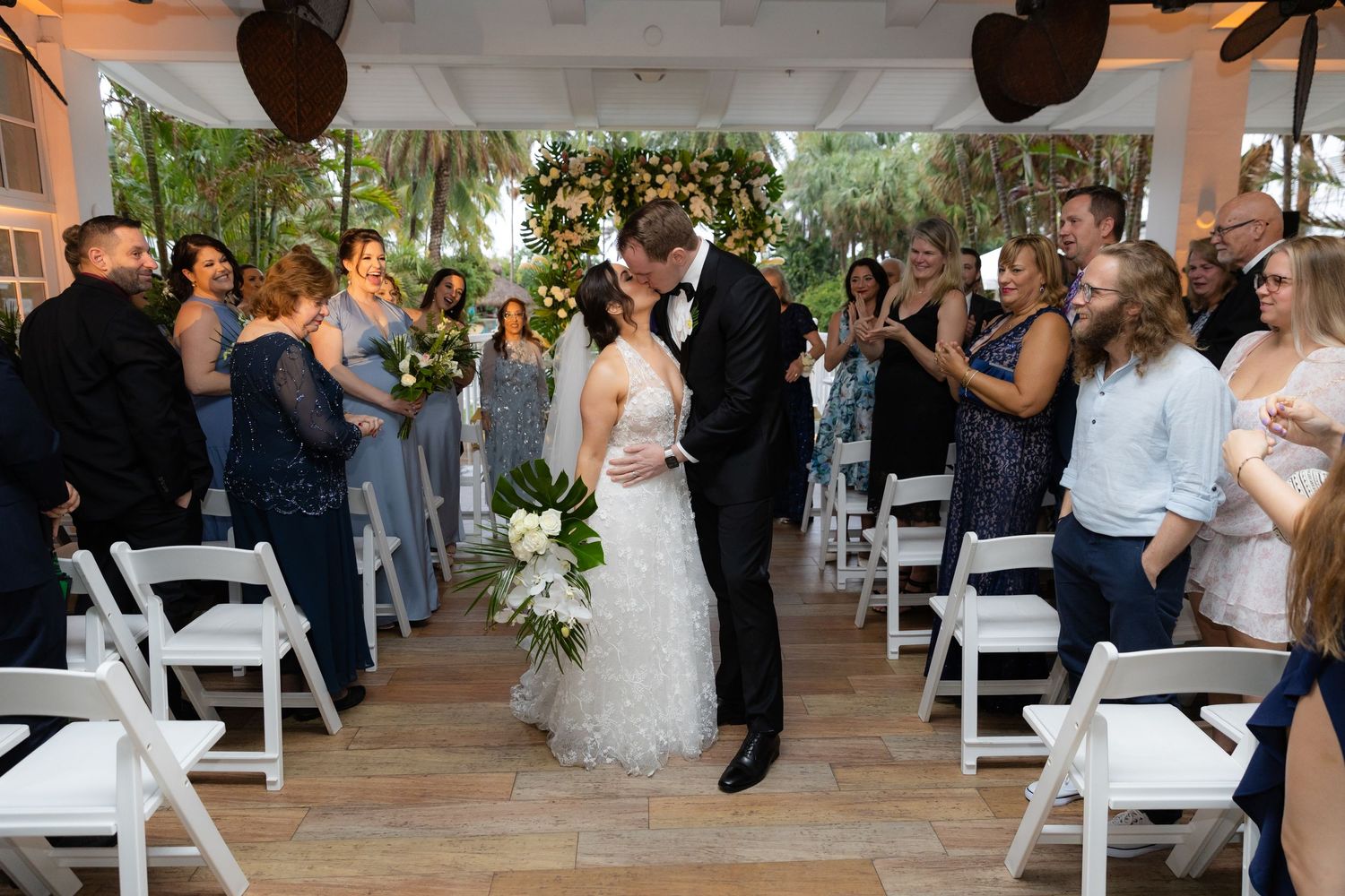 Wedding ceremony taking place outdoors with white chairs arranged in rows under trees.