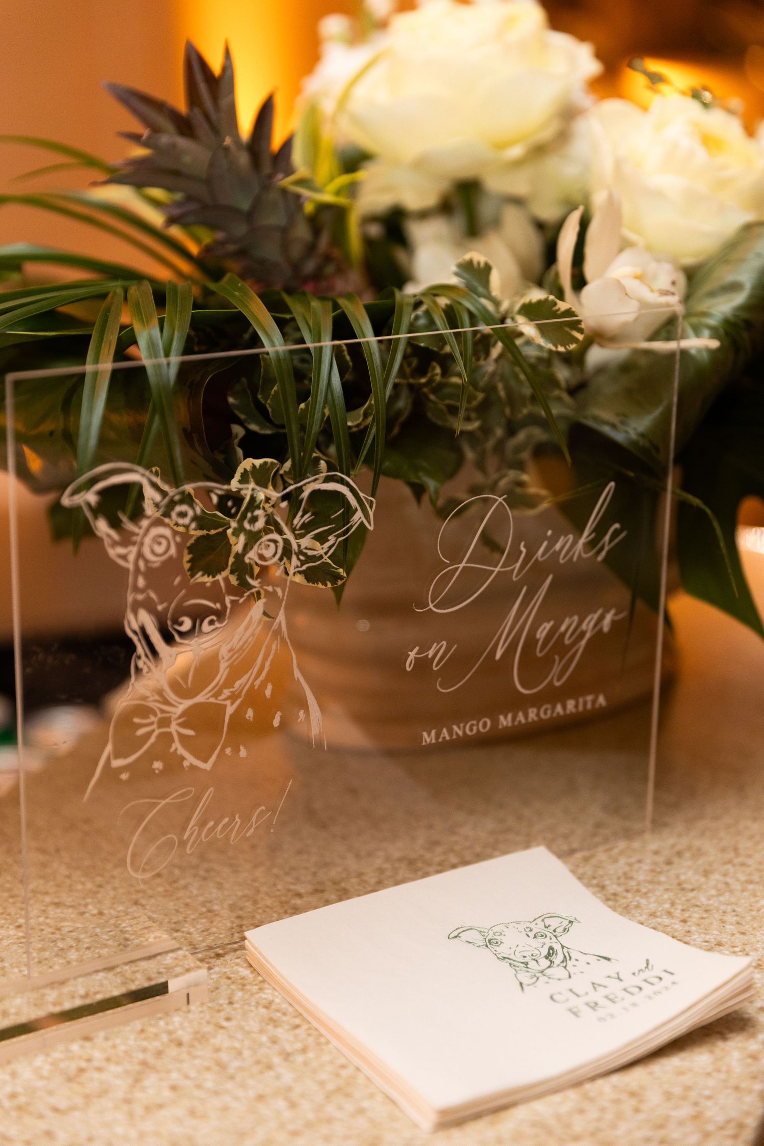 Clear acrylic wedding signage with elegant calligraphy displayed among floral arrangements.