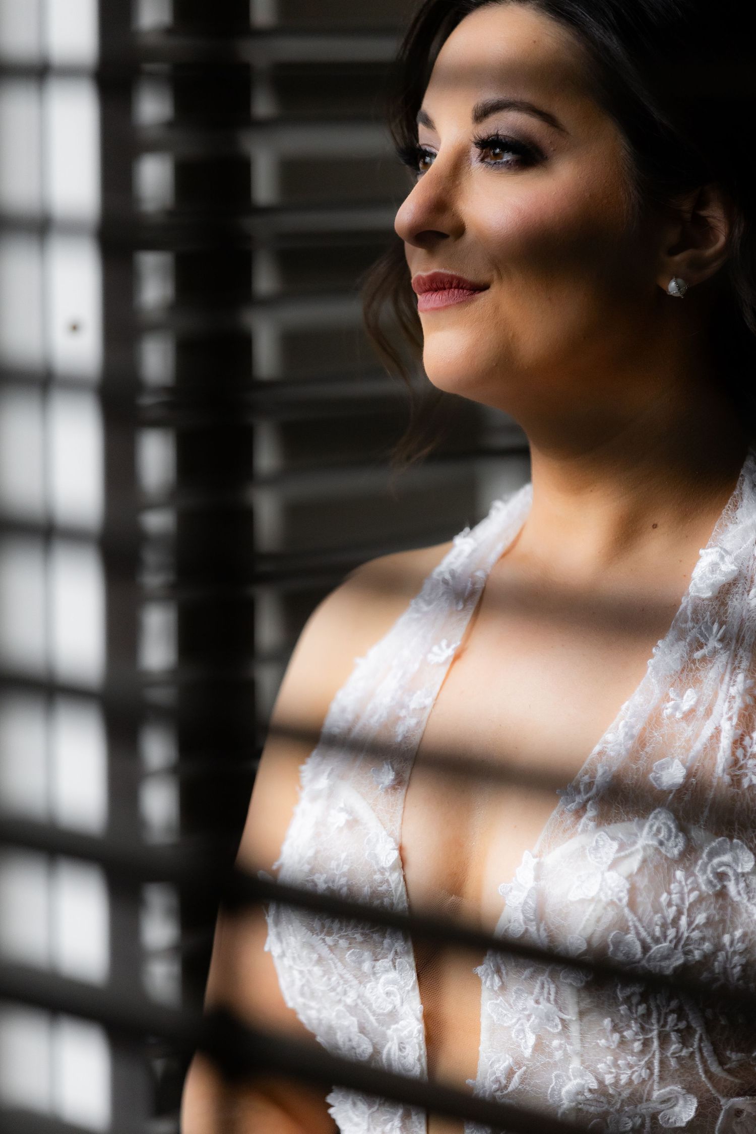 Artistic bridal portrait through window blinds creating striped light patterns on white lace dress.