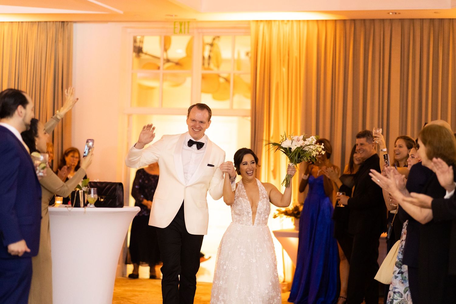 Wedding guests dance and celebrate at an elegant reception with warm amber lighting and white decor.