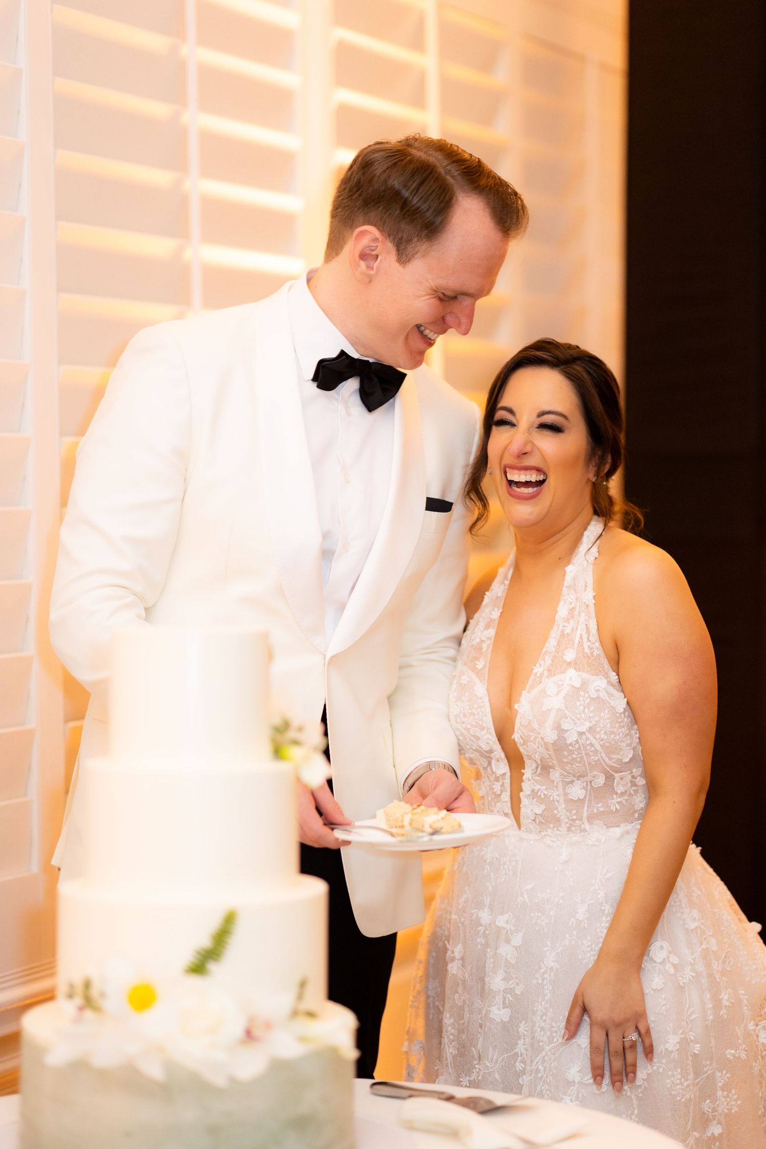 Happy couple laughs while cutting their tiered white wedding cake during their reception celebration.