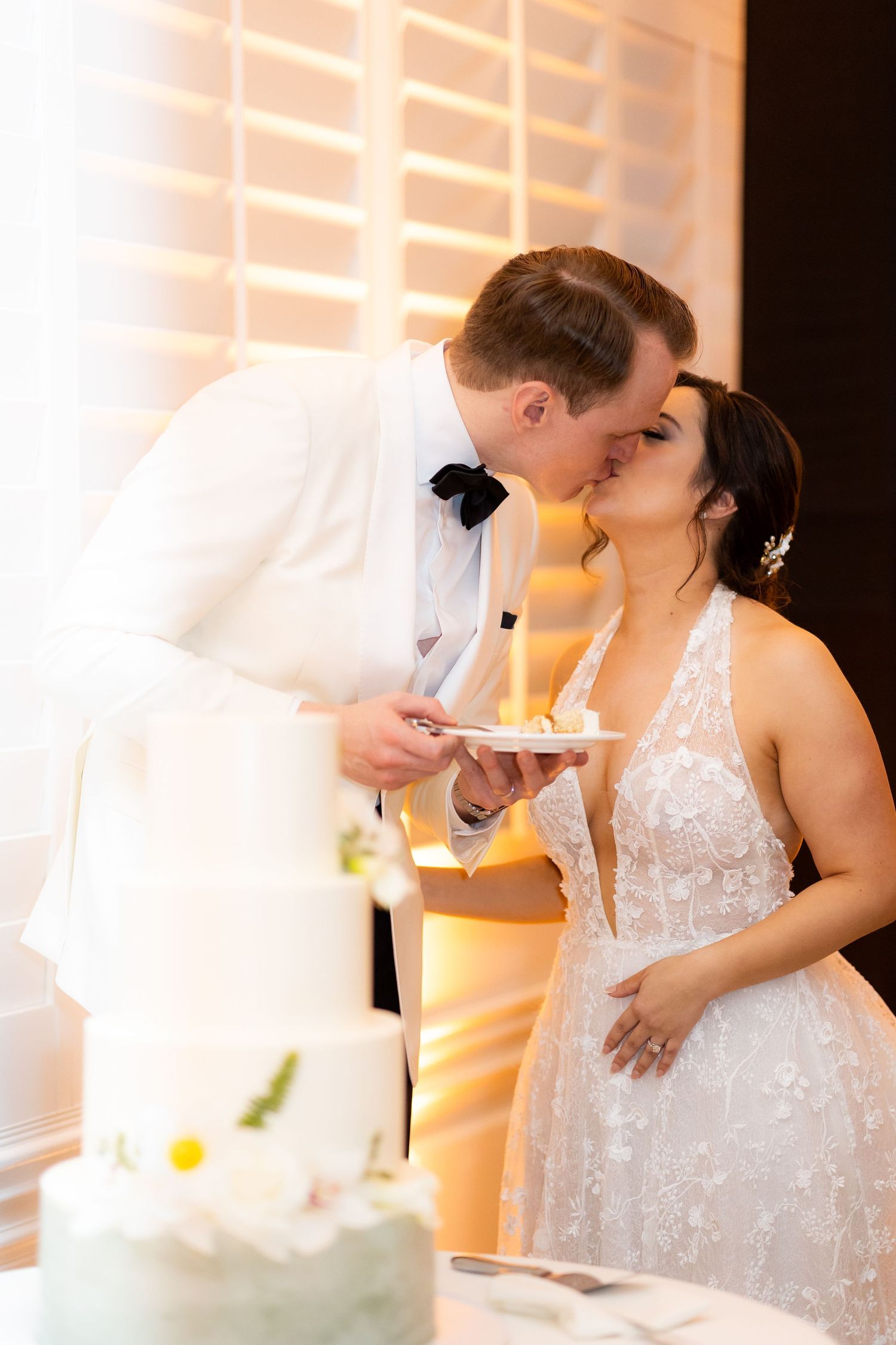Bride in sparkly dress and groom in white tuxedo share cake during wedding reception.