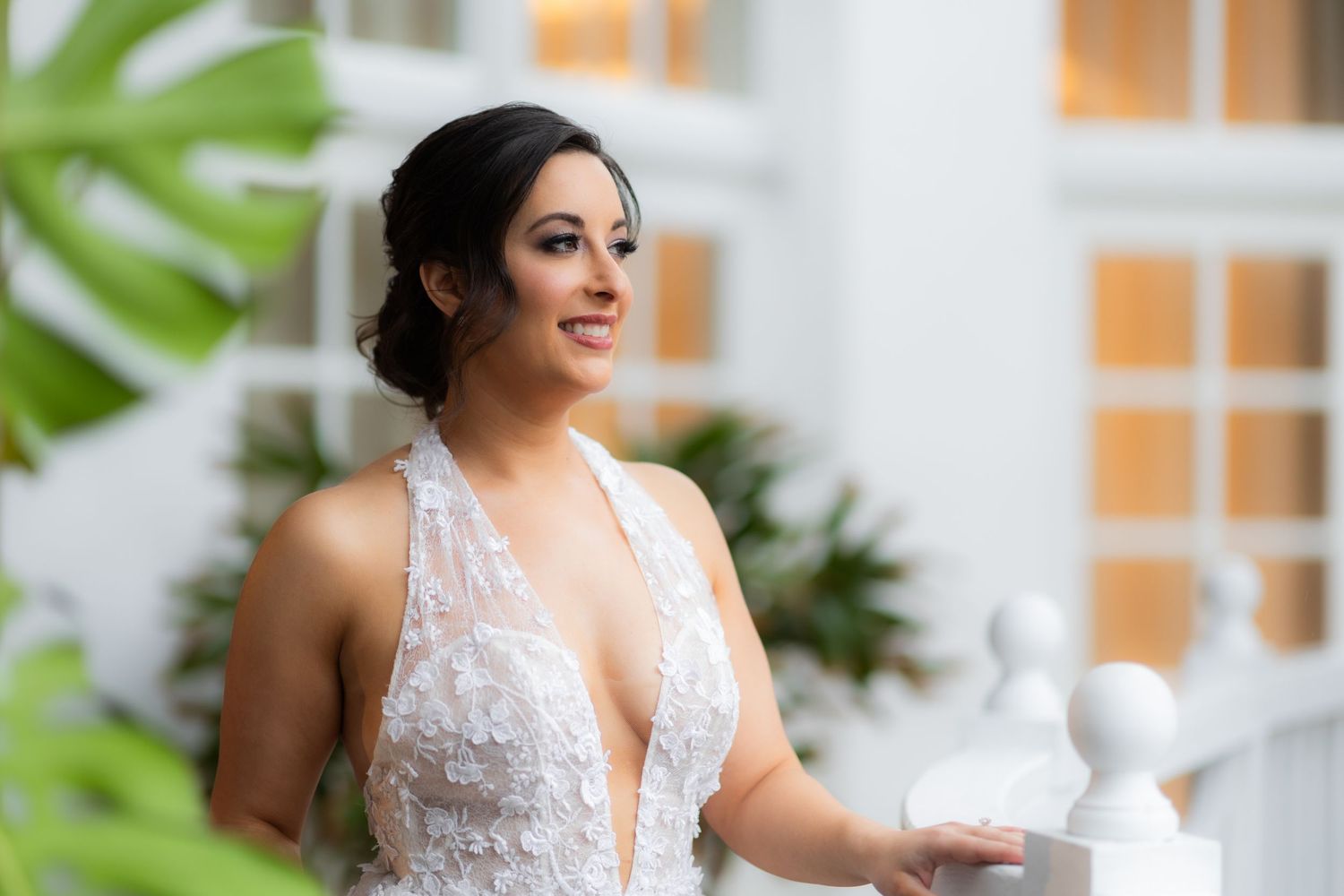 Bride in delicate lace V-neck gown poses on tropical veranda with palm trees in background.