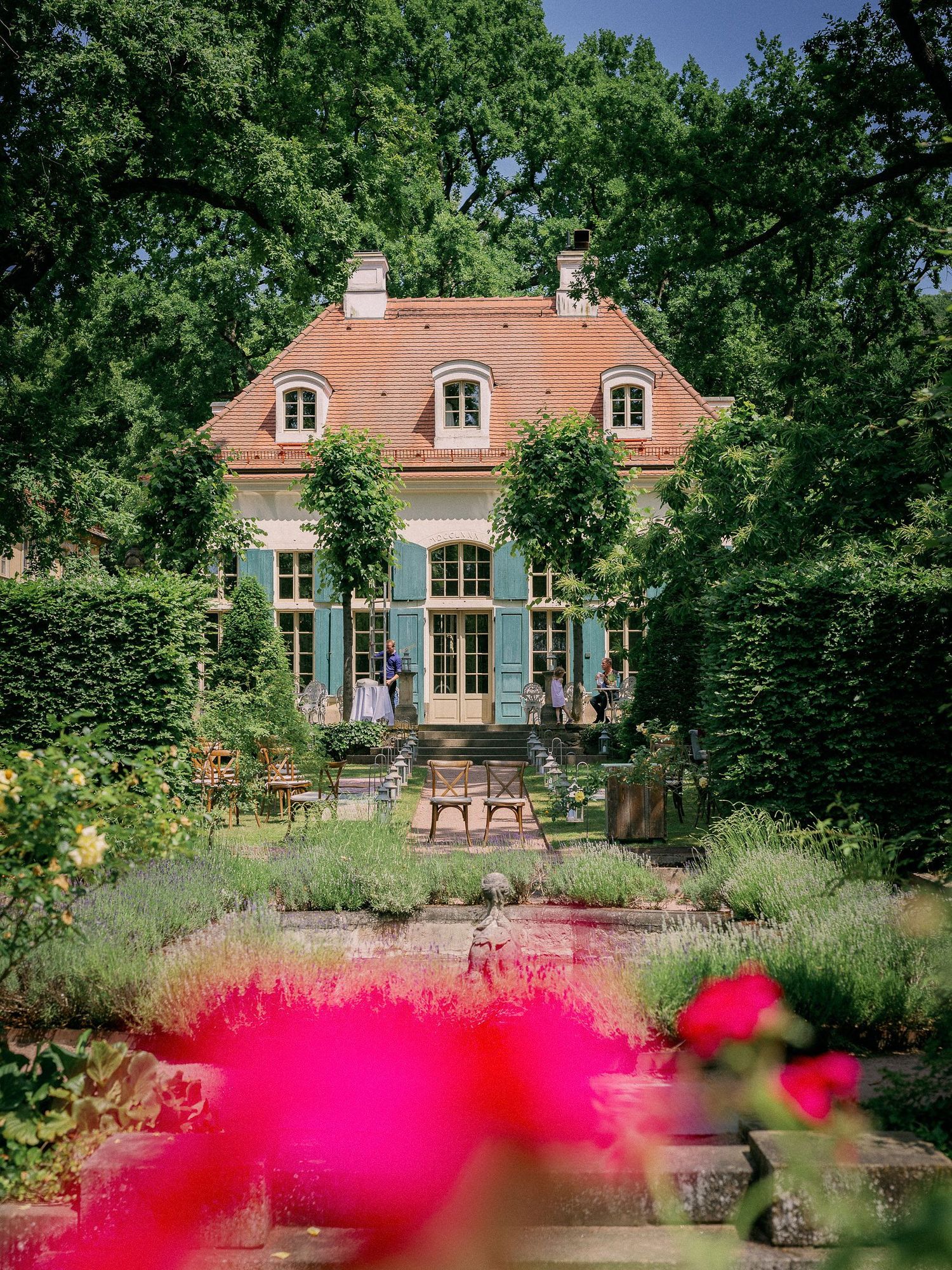 Charming mansion with blue shutters and formal garden featuring vibrant red flowers in the foreground.