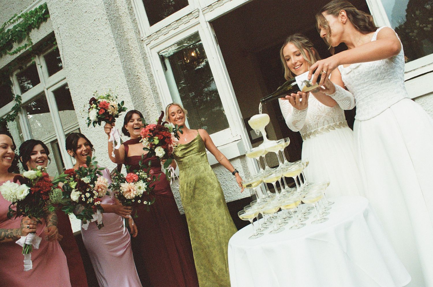 Bride and groom cutting white wedding cake while bridesmaids in pink dresses holding bouquets celebrate joyfully.