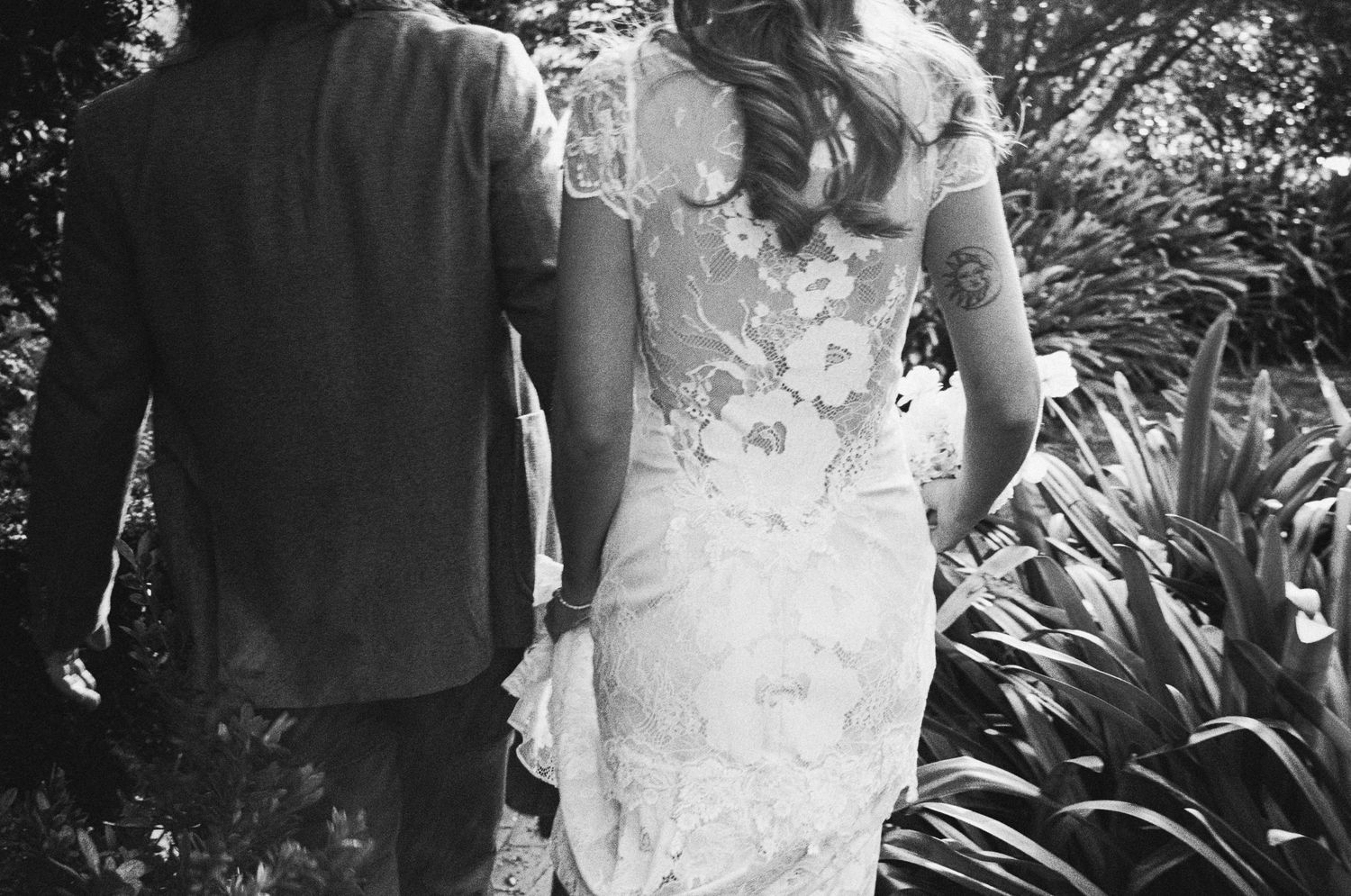 Black and white photo of bride in lace wedding dress holding hands with groom walking through garden.