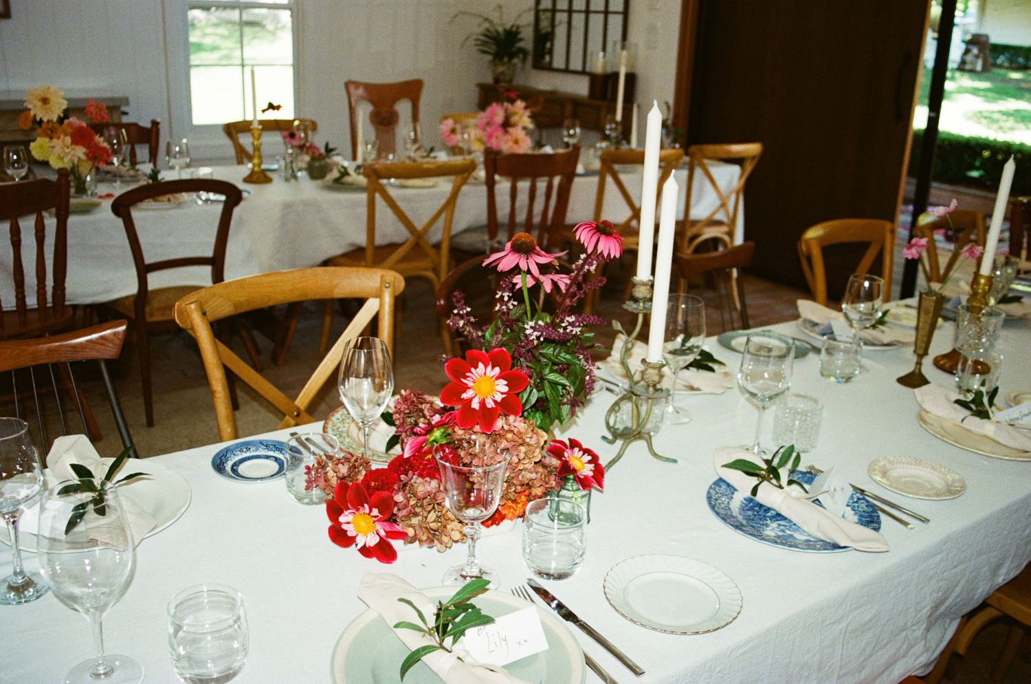 Elegant wedding reception table set with blue plates, white linens, red gerbera daisy centerpieces, and tall white candles.