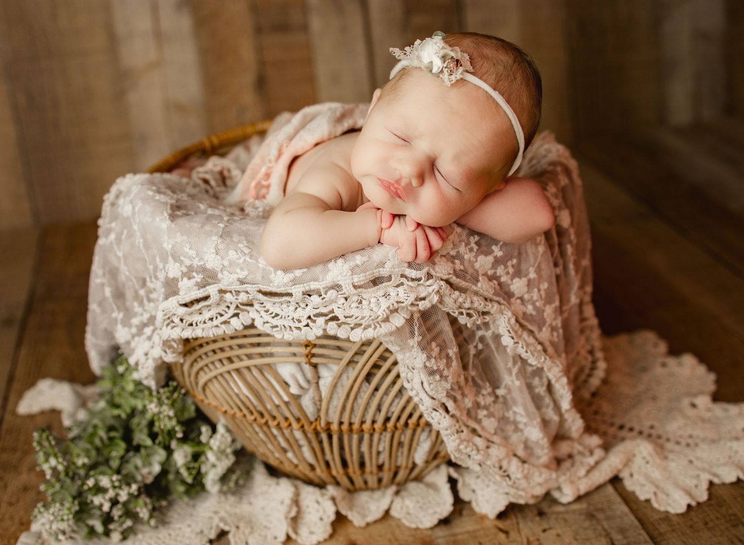 Sleeping newborn in lace wrap posed in rustic basket with floral accents.