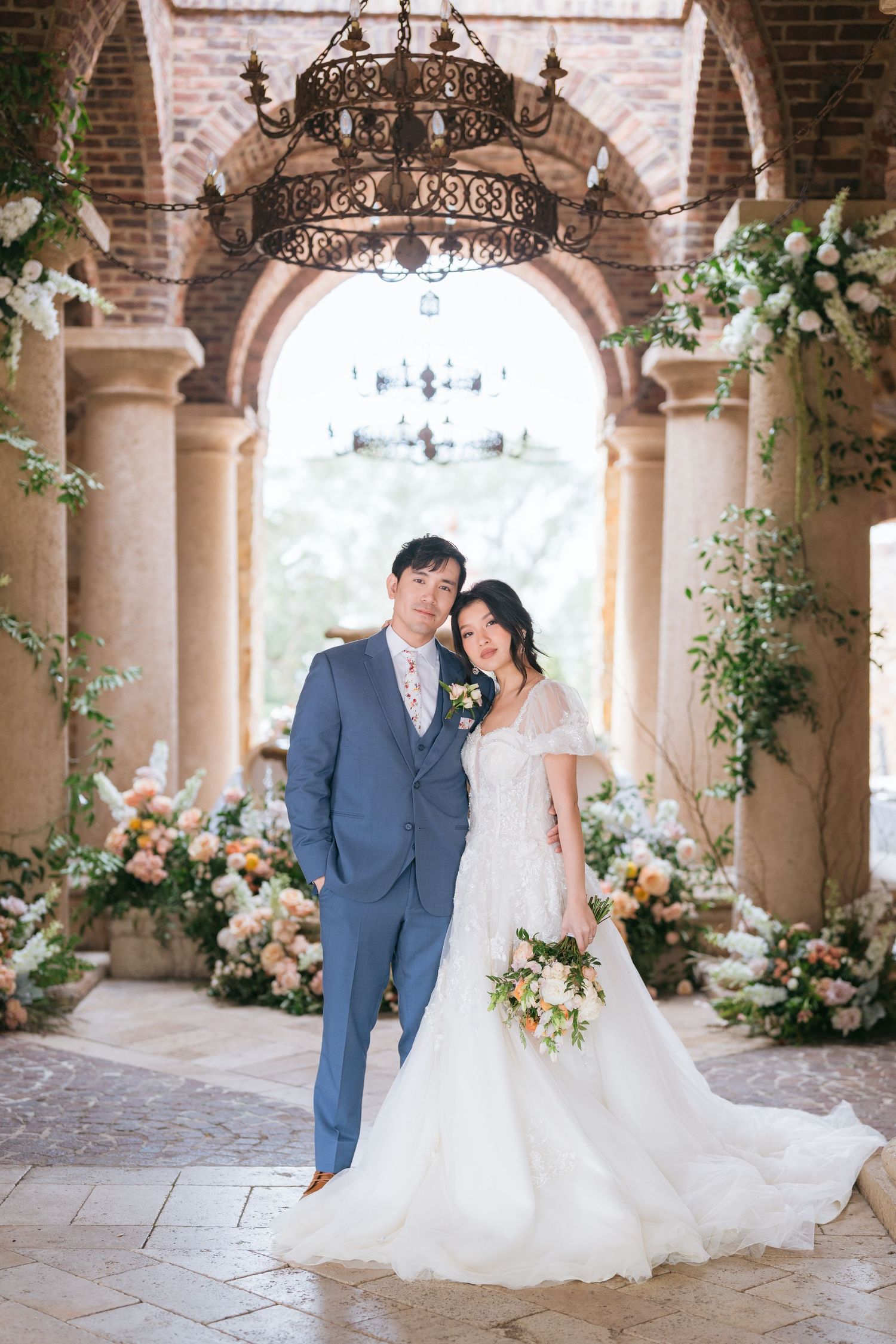 Elegant wedding scene in a stone colonnade decorated with floral arrangements and chandeliers.