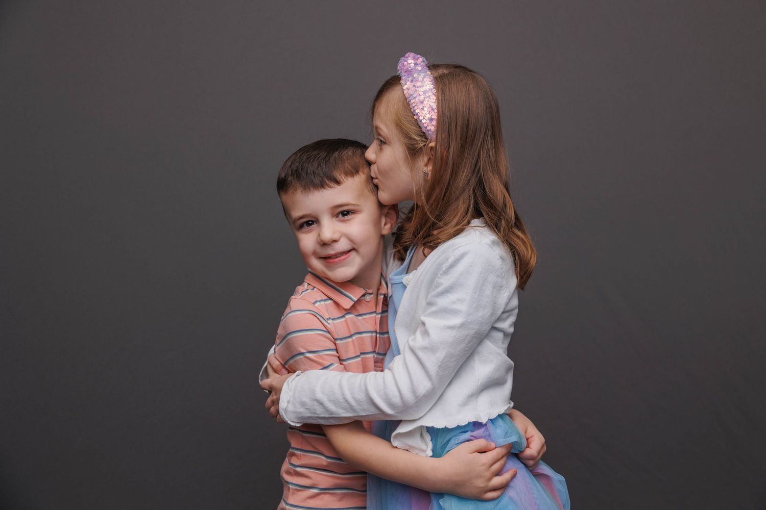 Two children share a sweet embrace against a dark background while wearing casual clothes.