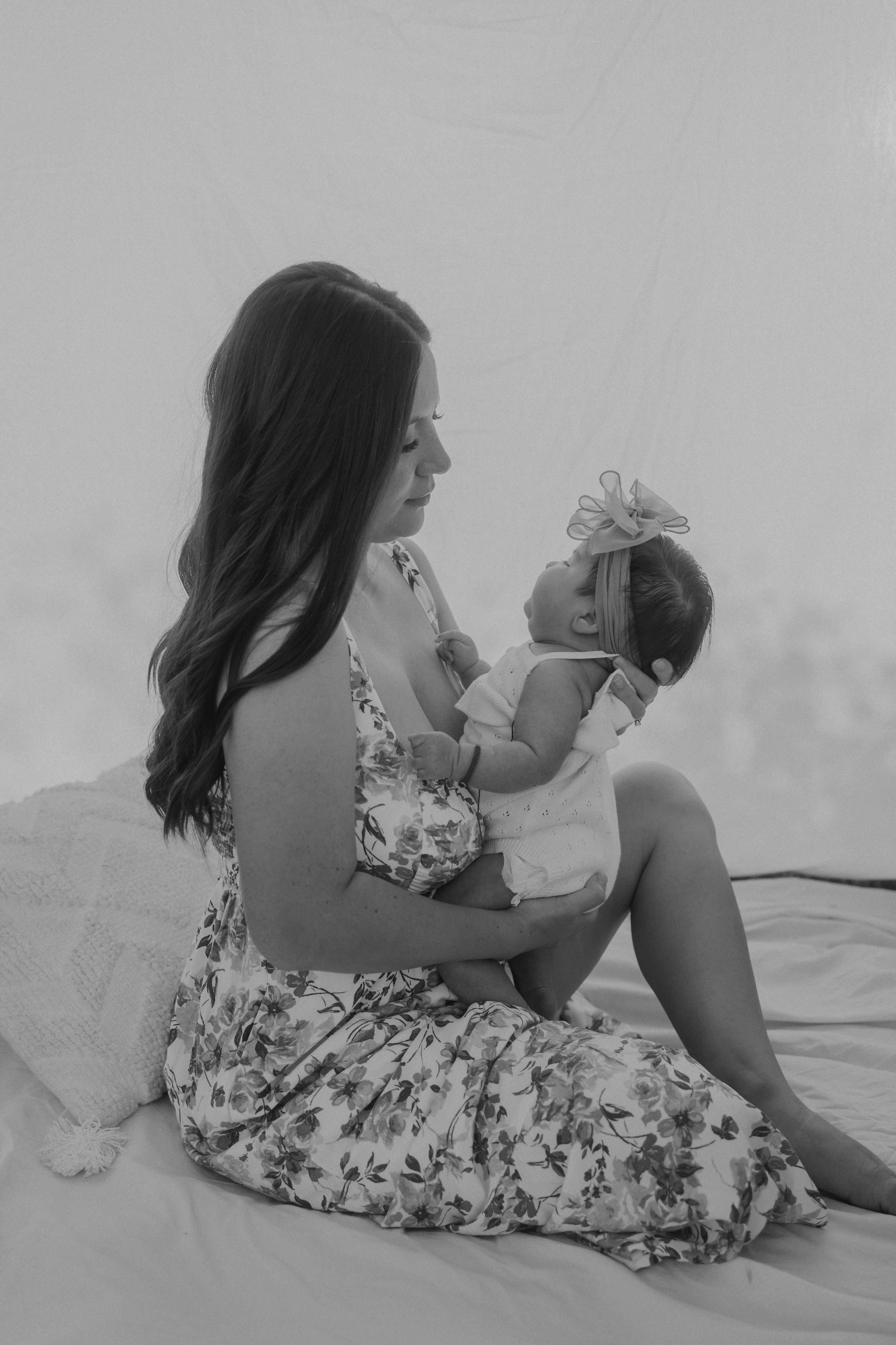 A woman in a floral dress gazes affectionately at a baby wearing a white outfit and a bow headband.