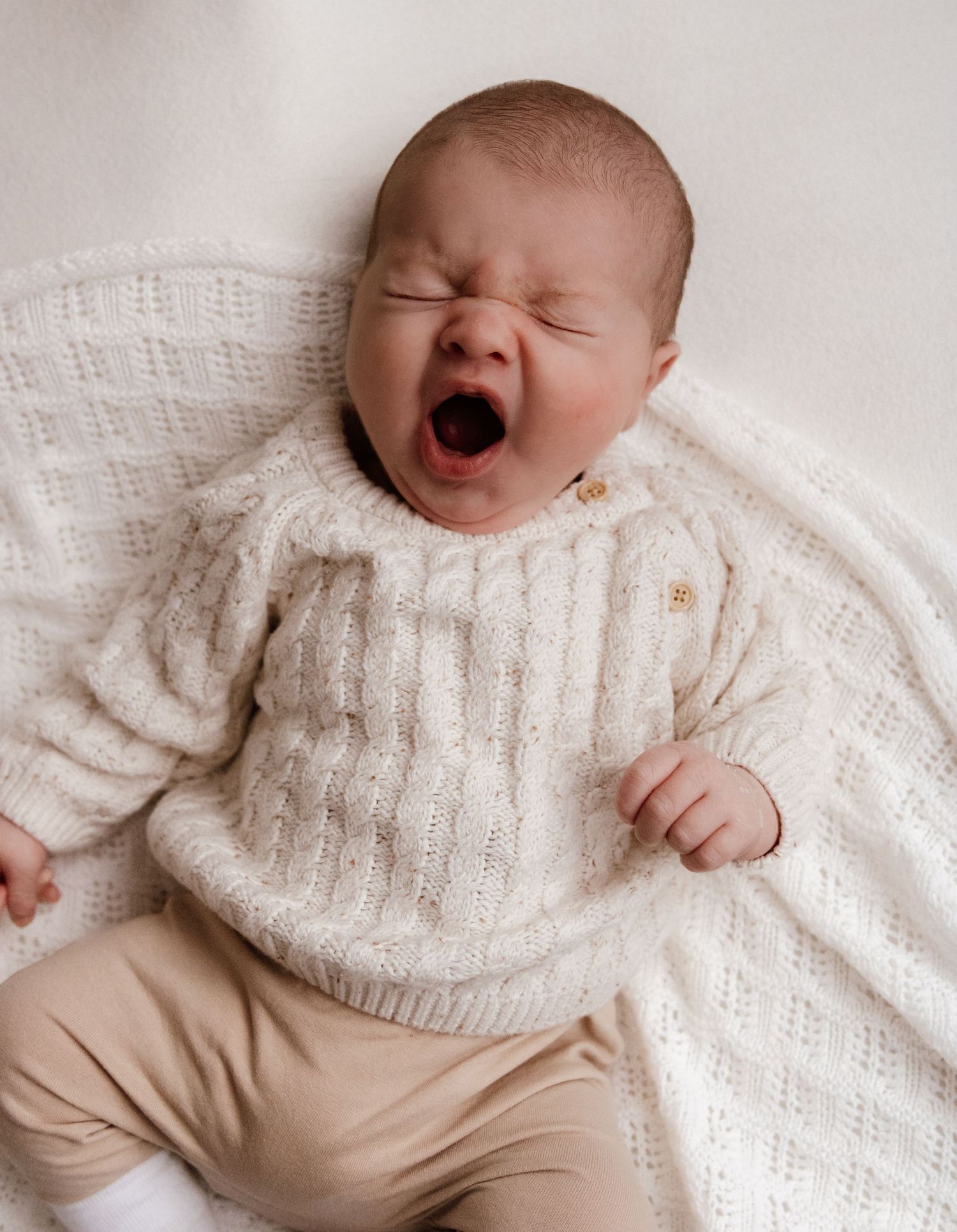 Newborn baby yawning while wearing cable knit sweater on white background.