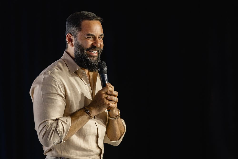 Speaker in beige shirt presenting on dark stage with microphone.