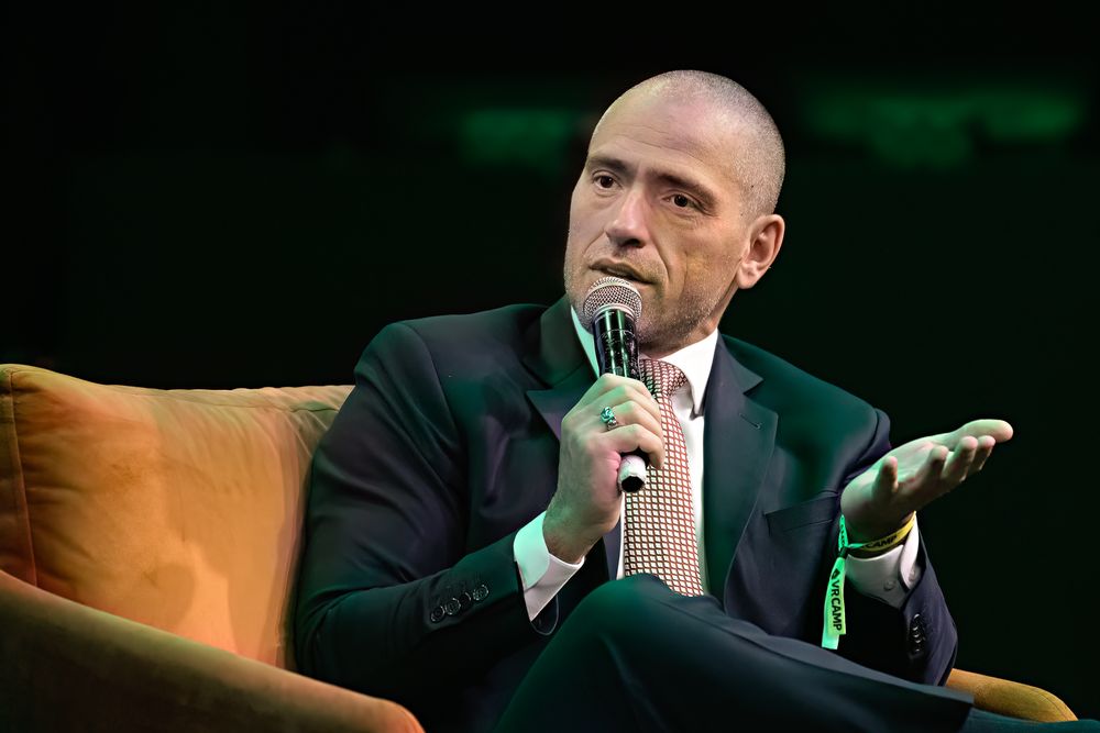 Business speaker in dark suit presenting on stage with dramatic lighting.