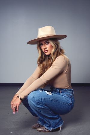 Model in beige top and jeans poses with wide-brim hat against gray backdrop.