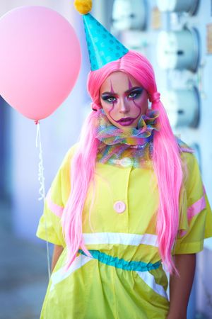 Colorful costume portrait with neon pink wig, party hat and yellow dress holding pink balloon.