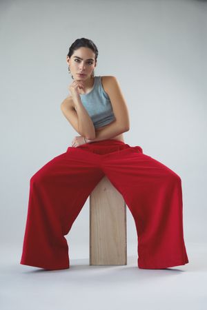 Fashion model in gray top and flowing red pants poses artistically on wooden block against white backdrop.