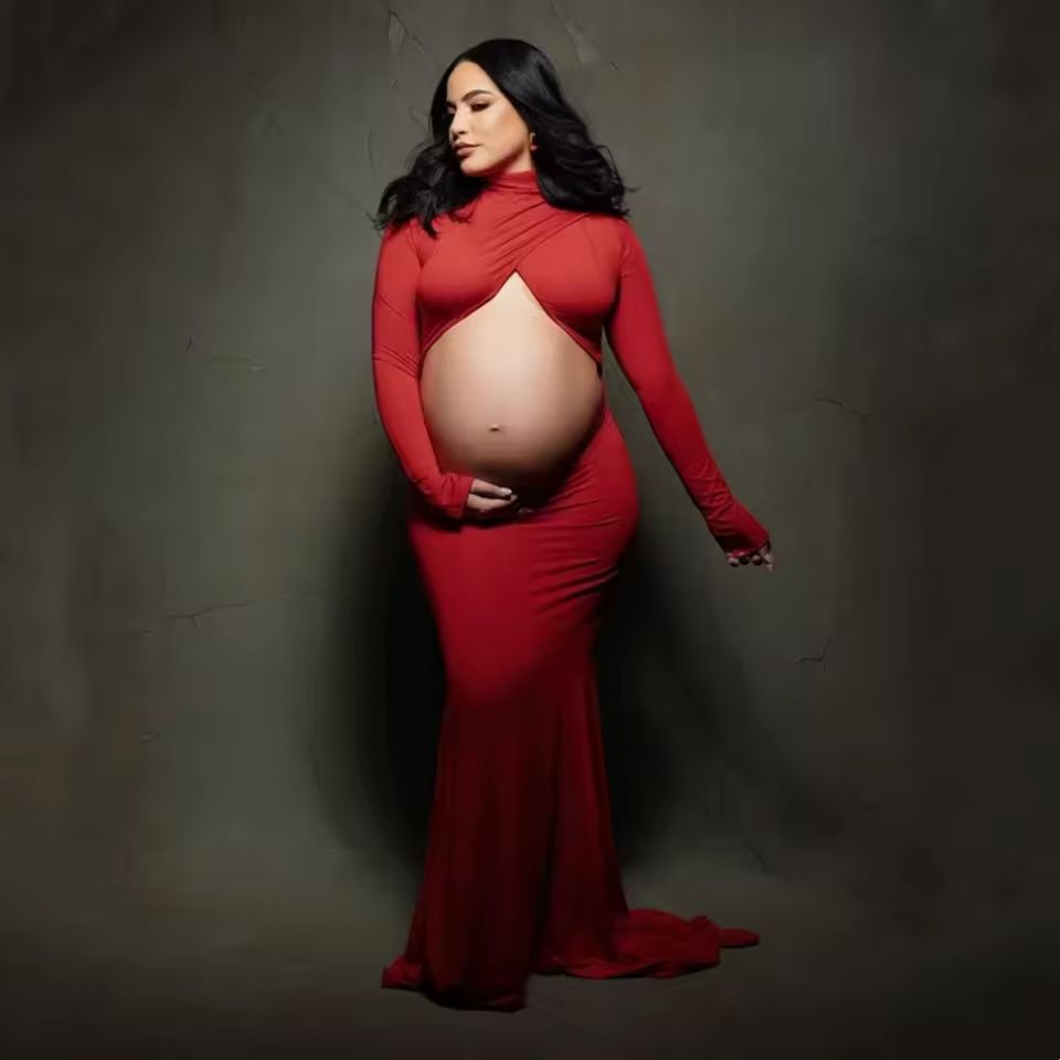 A woman in a flowing red maternity dress poses dramatically against a dark background during a photoshoot.