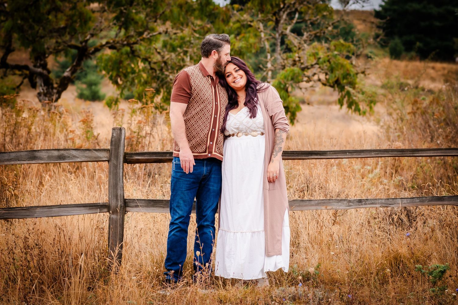 A couple in casual western attire stands together by a wooden fence in a golden autumn field at sunset.