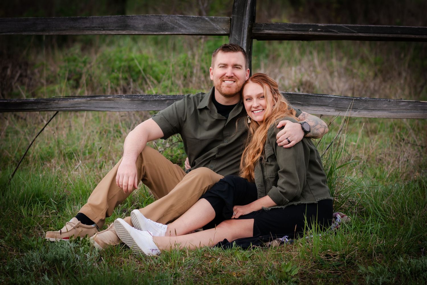 Couple in casual outdoor wear sitting together on grass near rustic wooden fence.