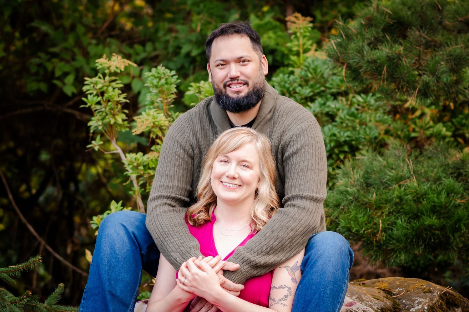 A couple sits together outdoors surrounded by lush greenery during a casual photo session.