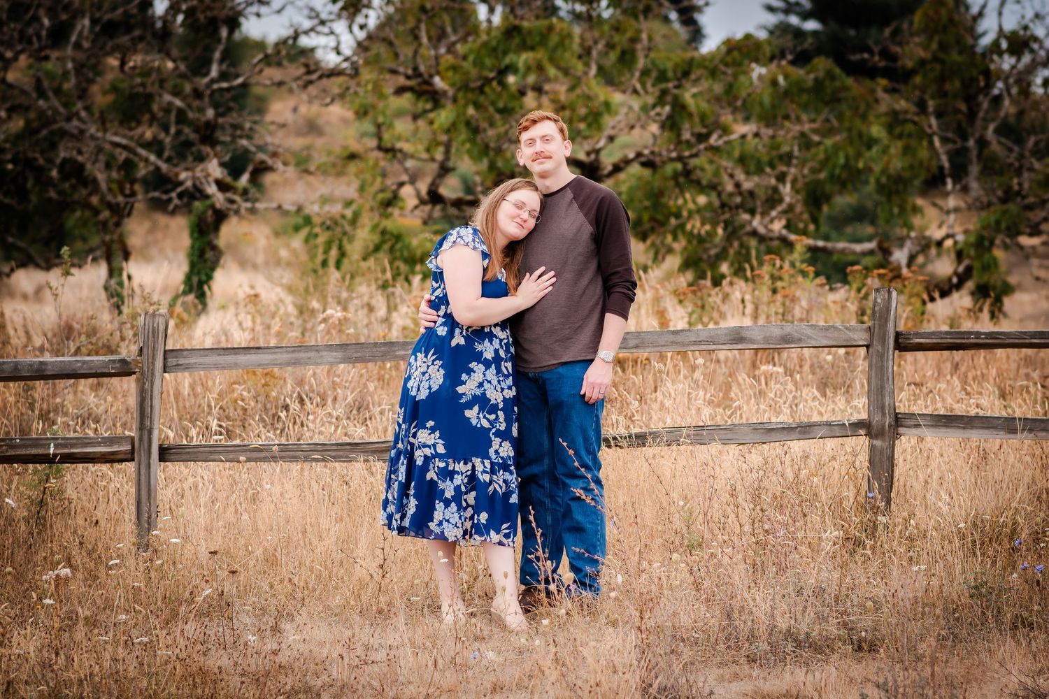 Couple embraces and smiles together on a nature path with wooden fence and dry grass in background.