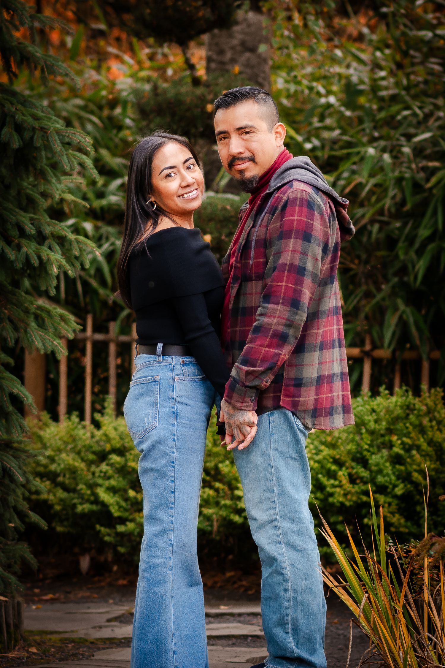 Couple in casual outfits posing together on garden path with evergreen backdrop.