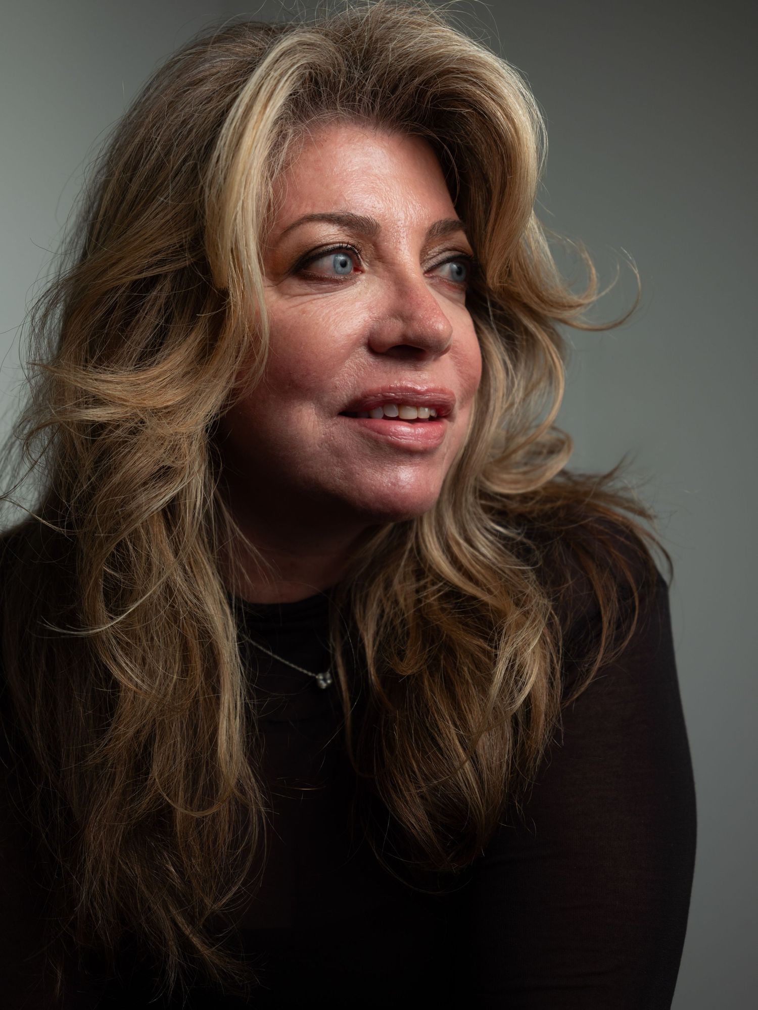 A portrait photo featuring someone with voluminous honey blonde hair wearing a black top against a dark background.