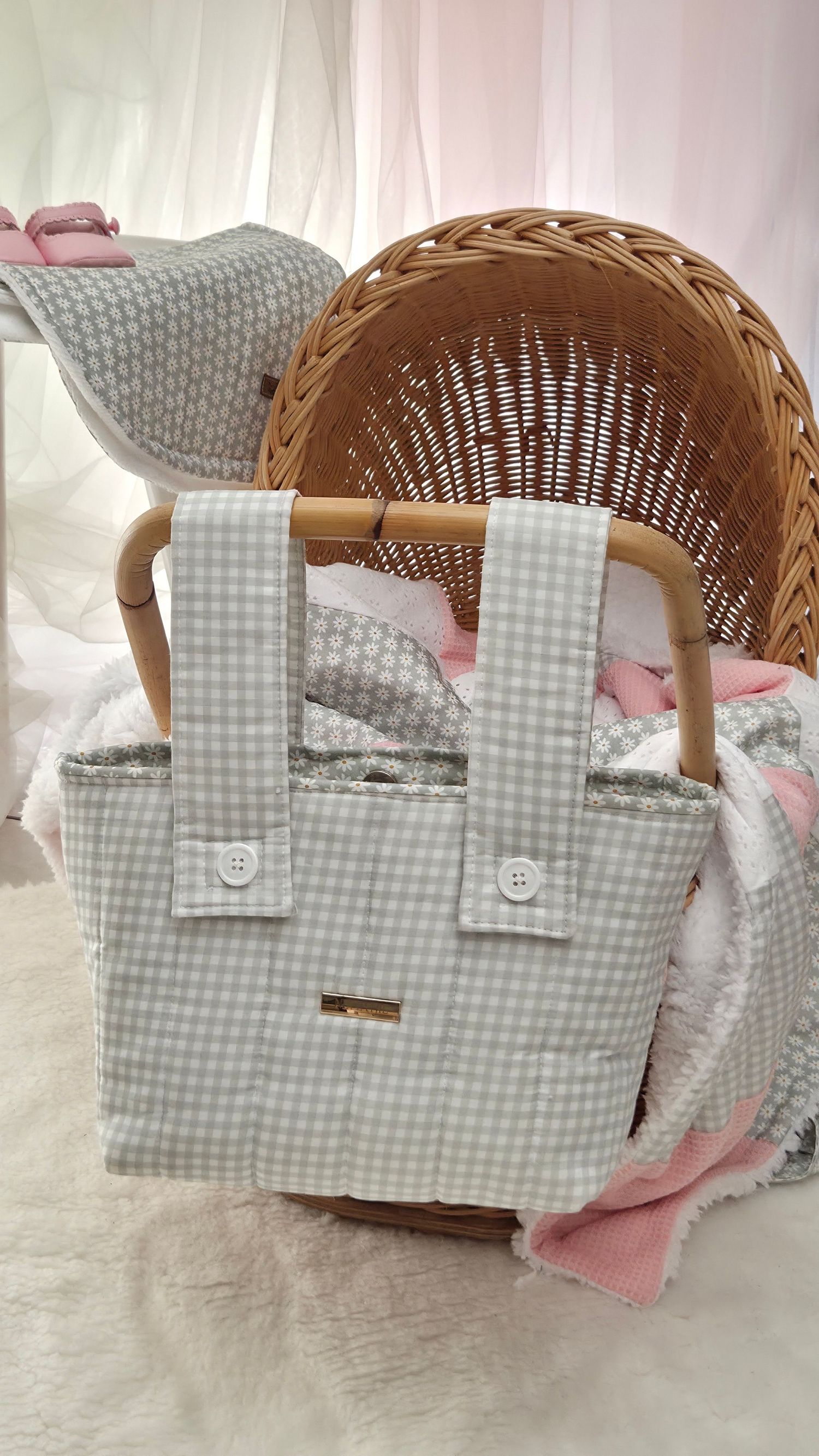 Gray gingham diaper bag with white buttons leaning against a wicker bassinet with pink and white bedding.