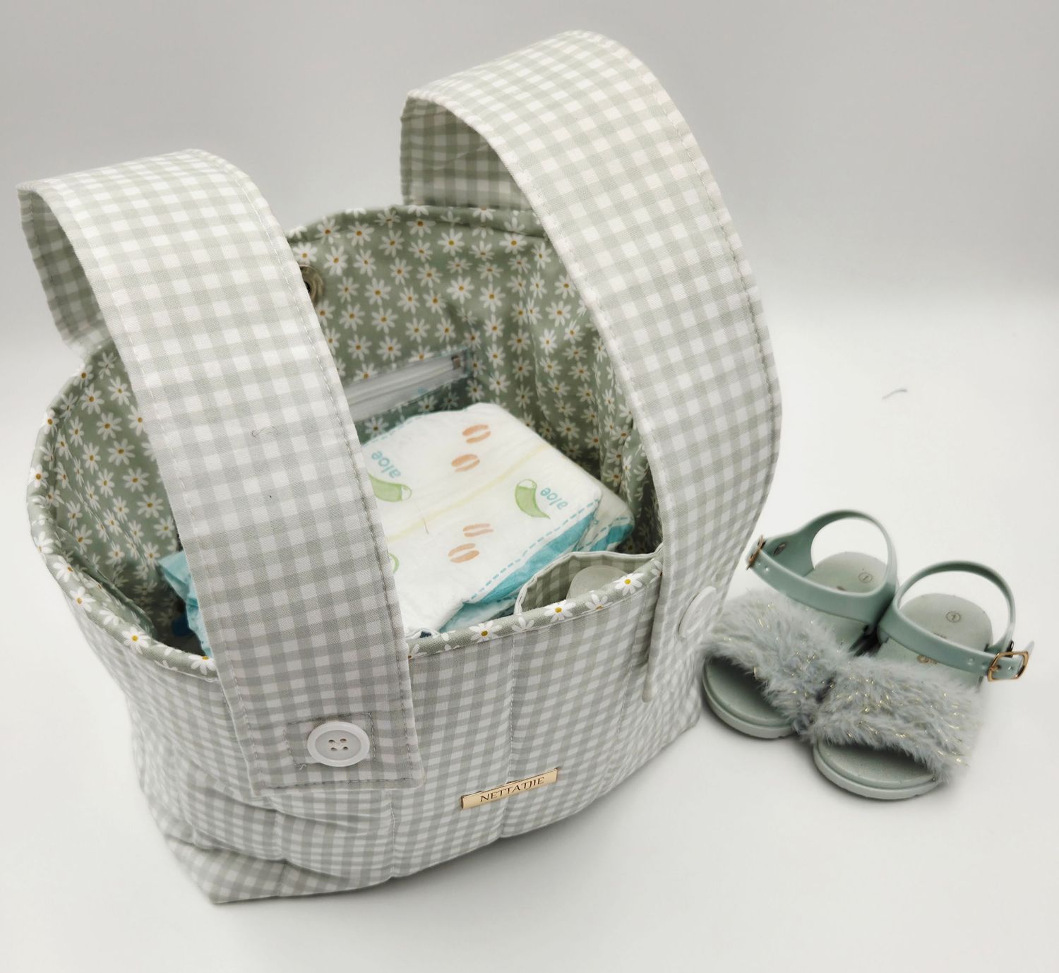 Gray gingham baby diaper bag with white button closure, featuring matching sandals and colorful patterned accessories inside.