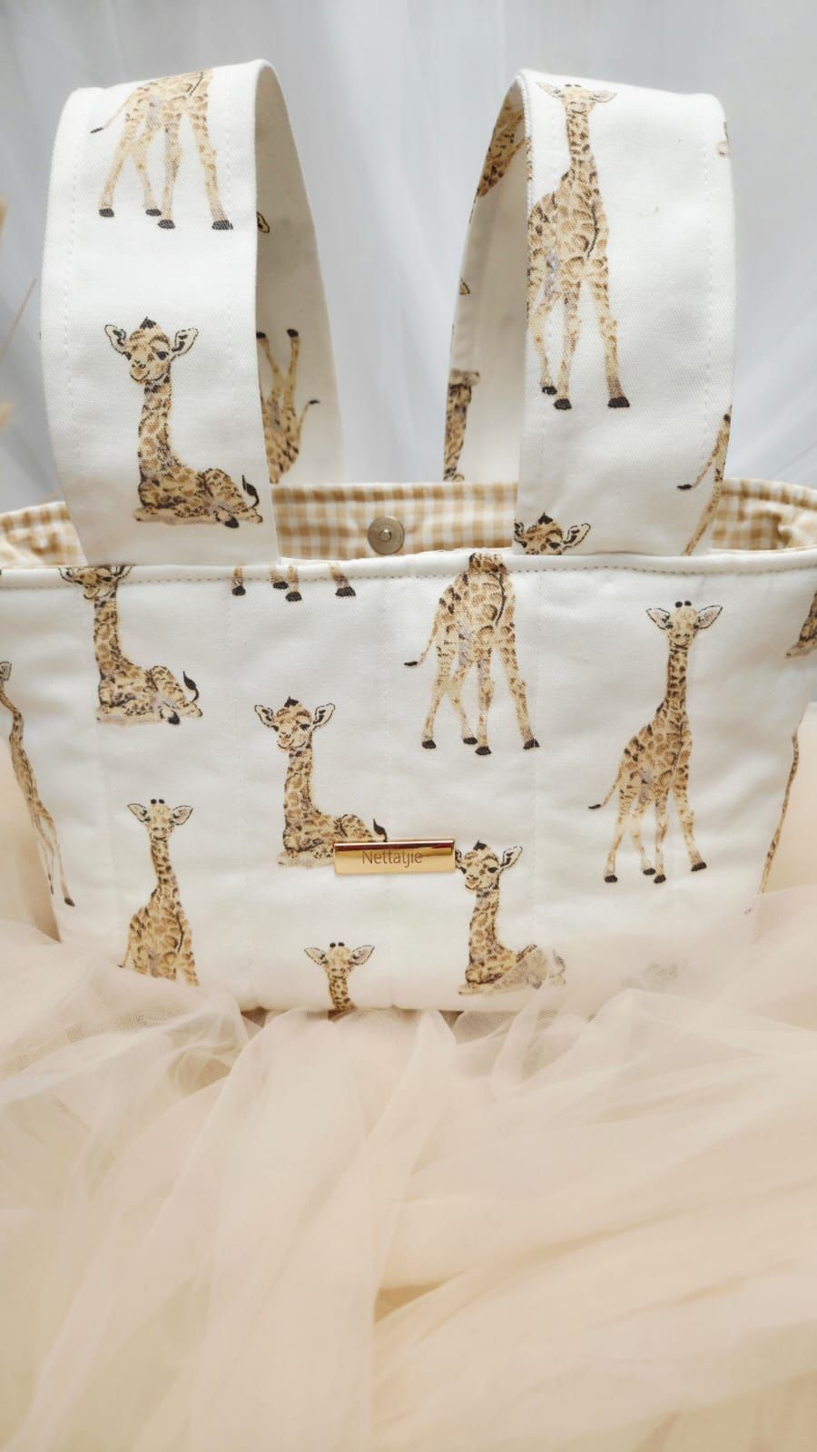 White fabric tote bag featuring adorable giraffe print pattern with tan gingham trim and handles.