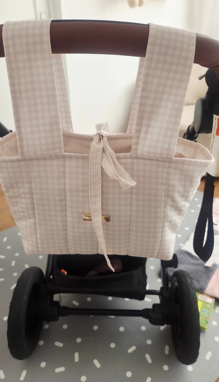Pink gingham diaper bag hanging on a baby stroller handle, featuring a bow tie detail and multiple pockets.