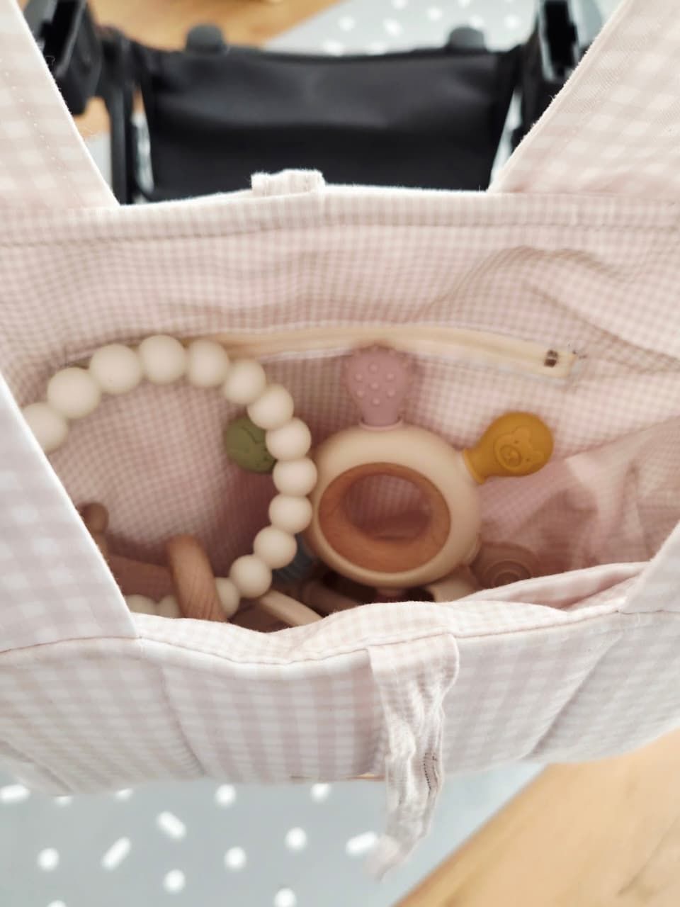 Pink gingham baby bag with wooden teething toys, silicone bead bracelet, and pastel rattles inside.