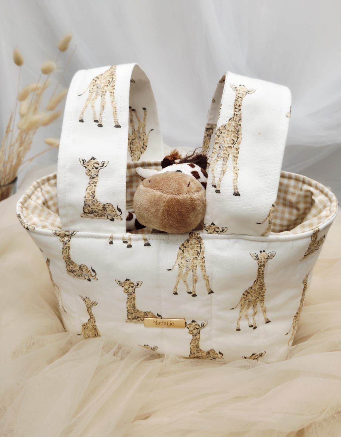 White fabric tote bag with giraffe print pattern and tan gingham lining, holding a plush giraffe toy.