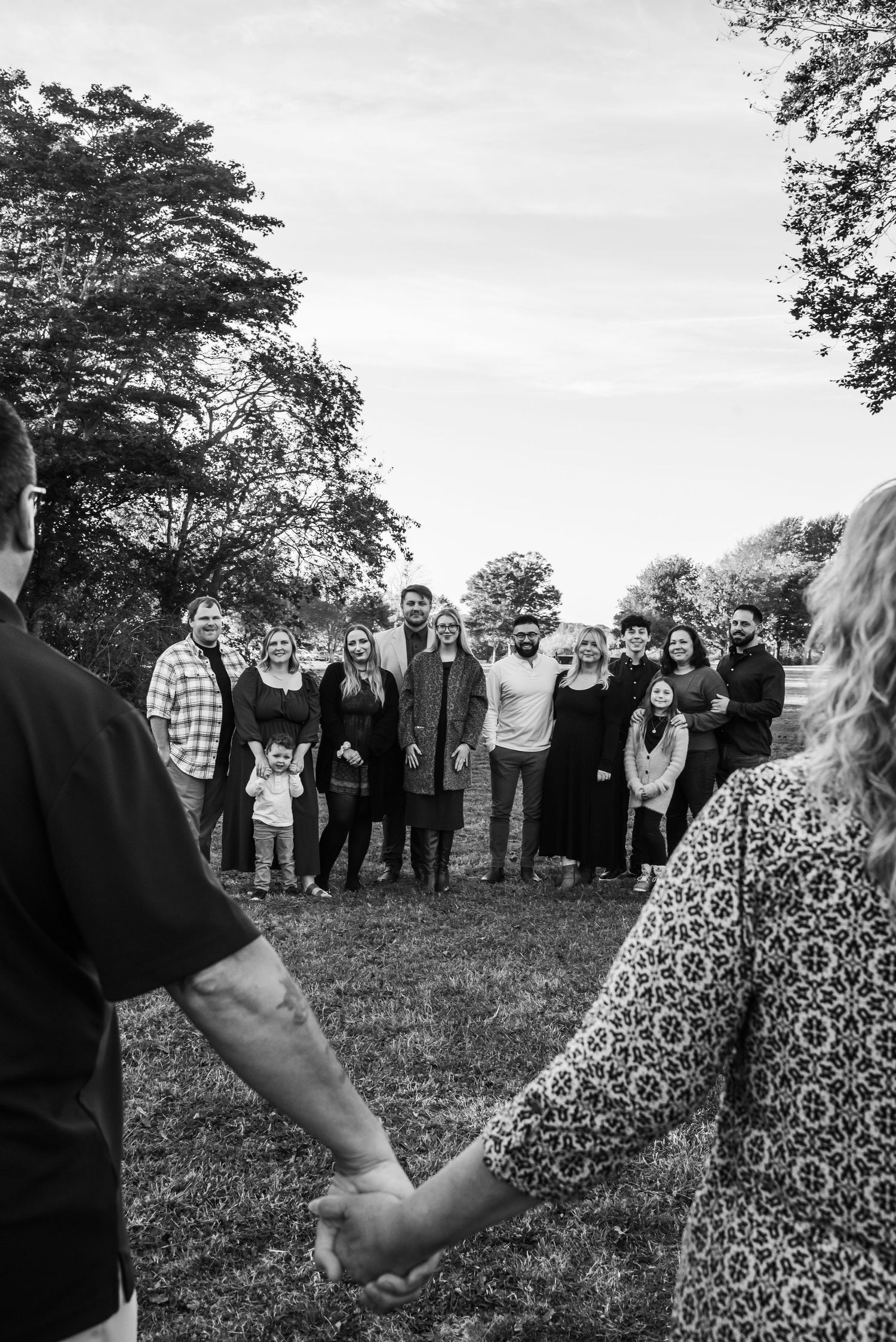 Black and white photo showing a group of people gathered outdoors under trees, with two joined hands visible in the foreground.